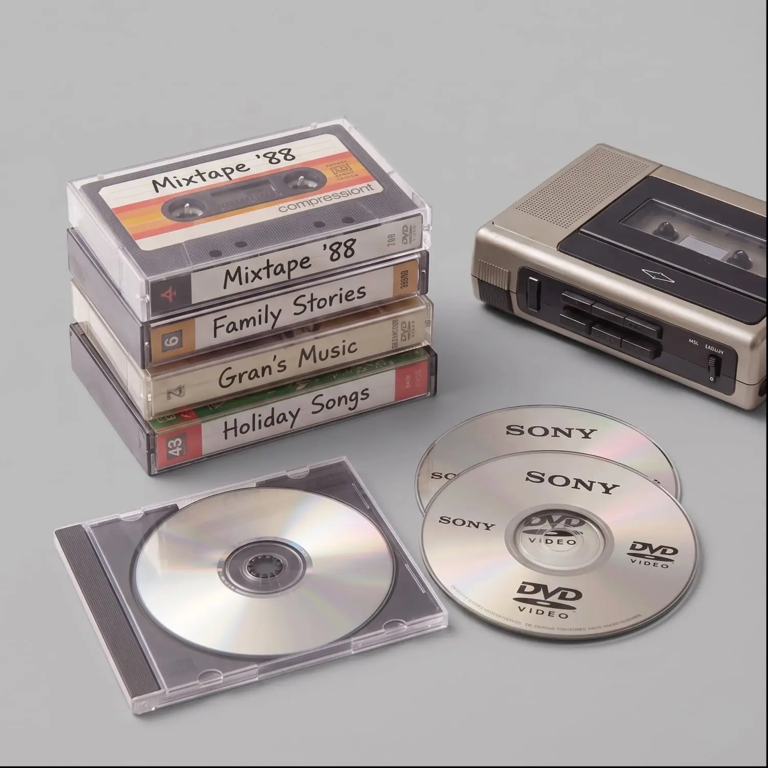 Stack of handwritten-titled cassette tapes next to a Sony portable player and several discs/CDs on a gray surface.